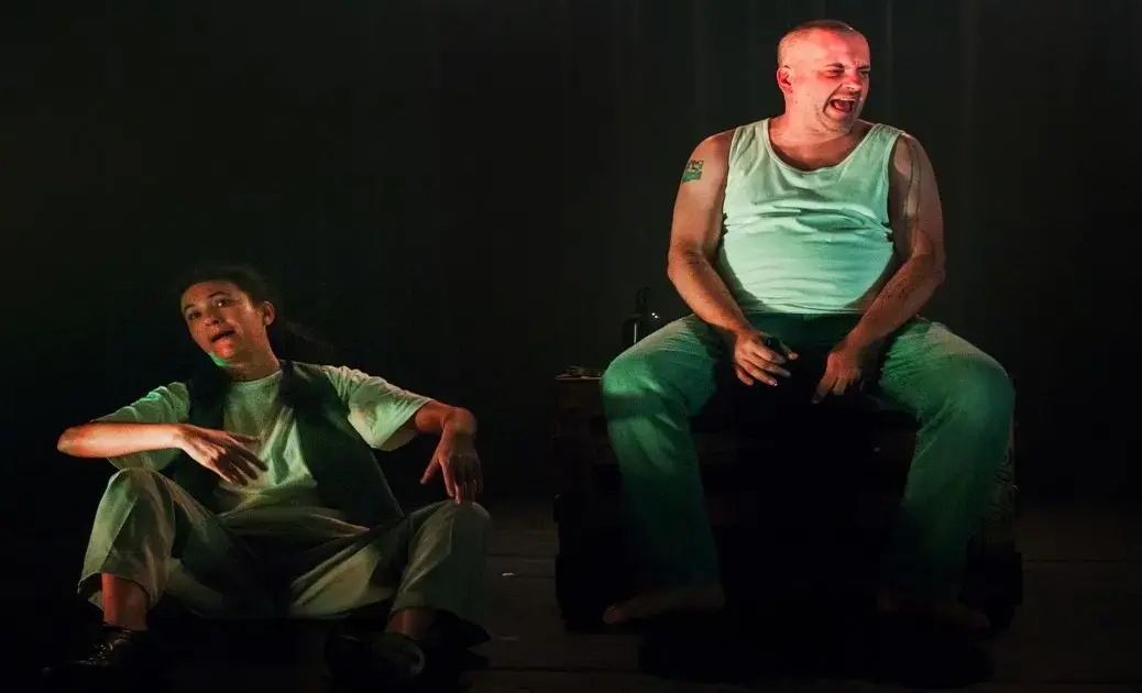 Duas pessoas em cena, uma sentada e outra em pé, aparentemente em um ambiente teatral.